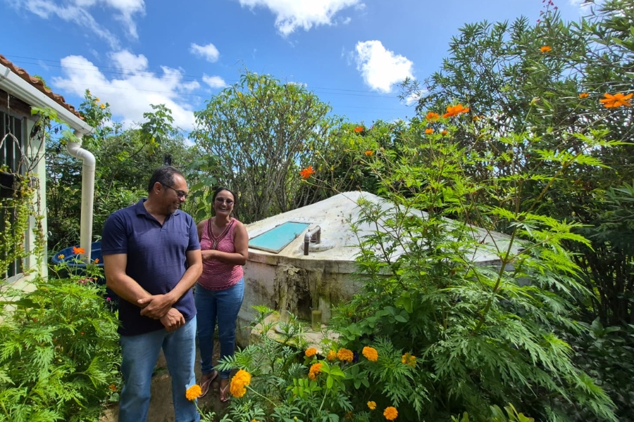 Um casal, Seu Zé e Dona Cilene, está em pé em um caminho estreito no quintal. O homem usa camisa polo azul e a mulher regata rosa. Ao lado deles, há uma grande cisterna de placas de concreto branca, cercada por arbustos e flores alaranjadas brilhantes. O céu está azul com nuvens brancas.