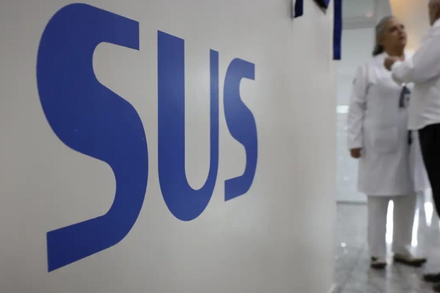 Sigla "SUS" escrita em letras azuis em uma parede branca no primeiro plano, com dois profissionais de jaleco branco desfocados ao fundo.
