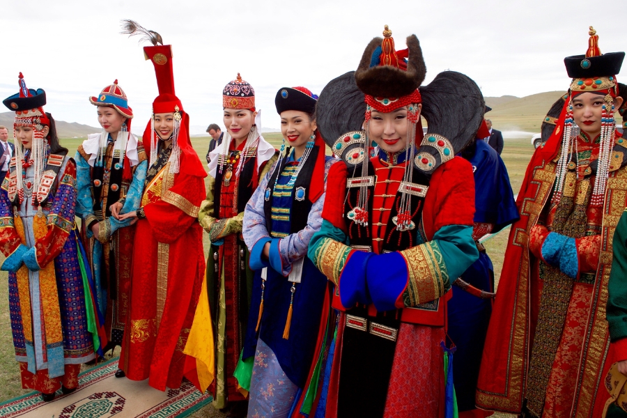 Grupo de mulheres posando ao ar livre vestindo trajes tradicionais mongóis coloridos, com túnicas detalhadas e adereços elaborados na cabeça.