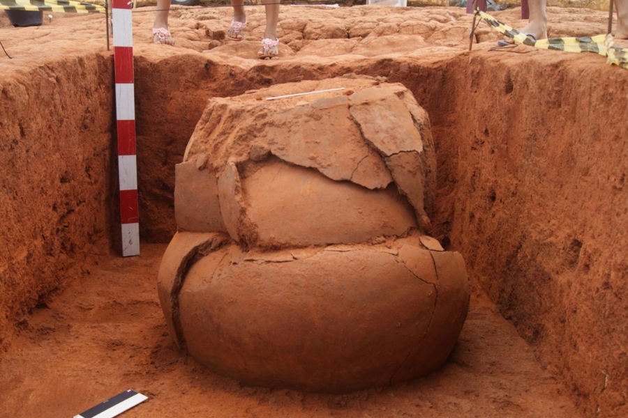 Sítio arqueológico exibe urna funerária de cerâmica fraturada em trincheira de terra vermelha. À esquerda, régua métrica preta e vermelha. Acima, pés humanos e fita de isolamento amarela.