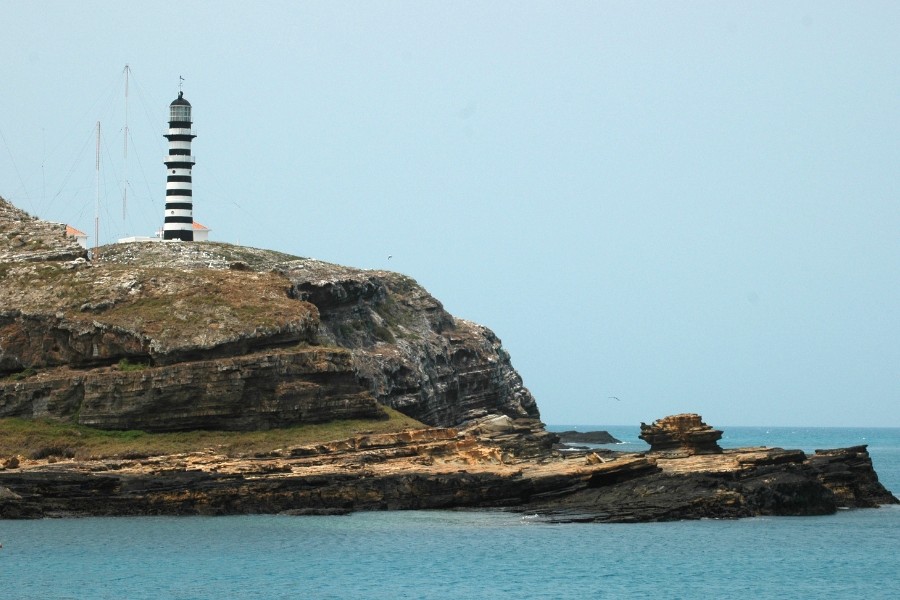 Fotografia horizontal de uma paisagem costeira com um farol no topo de uma ilha rochosa sob um céu azul-claro e limpo. No lado esquerdo, no ponto mais alto da elevação de terreno rochoso e acentuado, ergue-se um farol alto pintado com faixas horizontais intercaladas em preto e branco, com cúpula escura e uma pequena estrutura com telhado avermelhado na base, além de antenas ao fundo. O relevo é composto por rochas escuras dispostas em camadas horizontais visíveis, cobertas por uma vegetação baixa e rasteira em tons de verde e marrom. A base da falésia estende-se em direção ao centro e à direita até encontrar o mar de águas em tom azul-turquesa, onde formações rochosas rasas e escuras emergem perto da superfície