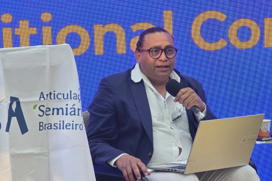 Em plano médio, um homem negro de pele clara, com cabelos escuros presos para trás e óculos de armação arredondada escura, fala ao microfone que segura com a mão esquerda enquanto olha para a frente. Ele veste uma camisa social branca com o colarinho aberto, um paletó azul-marinho e calças claras; em seu colo está um notebook prata aberto, sobre o qual ele apoia a mão direita no touchpad. À esquerda da imagem, em primeiro plano, aparece a quina de um pódio coberto por um pano branco com o logotipo "ASA" e a frase "Articulação Semiárido Brasileiro". Ao fundo, destaca-se um painel digital azul brilhante com as letras amarelas "itional Co" na parte superior