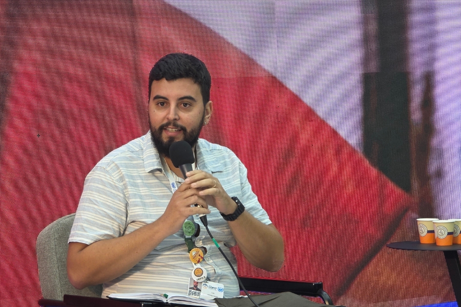 A fotografia, em plano médio, de um homem jovem de pele clara, cabelos pretos curtos e barba cheia, sentado enquanto se pronuncia durante o painel. Ele veste uma camisa de mangas curtas com listras horizontais sutis em tons de azul-claro e bege, e traz no peito uma fita de crachá decorada com botões e adesivos do evento. Segura com ambas as mãos um microfone preto próximo ao queixo, mantendo um olhar atento e um leve sorriso. Ele está sentado em uma poltrona cinza-claro, com um caderno aberto sobre o colo e uma pequena mesa lateral à direita onde descansam copos descartáveis. O fundo é composto por um painel de LED com textura quadriculada visível e tons avermelhados