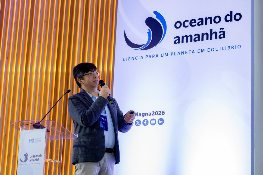 Fotografia horizontal em ambiente interno mostra um homem em pé, no lado esquerdo, realizando uma apresentação durante um evento. Ele é um homem adulto de pele clara, cabelos curtos e escuros com franja caindo levemente sobre a testa, e usa óculos de grau. Veste um blazer cinza-escuro, camisa de botão clara por baixo, calça bege e carrega um crachá pendurado no pescoço. Ele segura um microfone preto próximo à boca com a mão esquerda e um passador de slides na mão direita, olhando para a direita com a boca levemente aberta. À sua frente há um púlpito de acrílico transparente com um microfone fixo de haste fina e o logotipo do evento impresso na parte frontal. Ao fundo, no lado esquerdo, há um painel decorativo com ripas de madeira verticais em tom alaranjado iluminadas; no lado direito, há um grande painel branco onde se destaca o logotipo de uma onda em tom azul-escuro com o texto "oceano do amanhã", seguido pelo slogan "CIÊNCIA PARA UM PLANETA EM EQUILÍBRIO", além do texto "Magna2026" e ícones de redes sociais