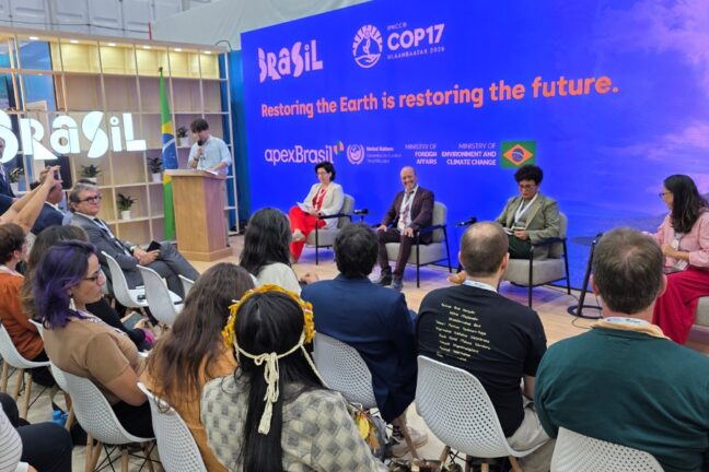 Fotografia de um painel de discussão em um estande, há um painel azul vibrante com o logotipo estilizado "Brasil", a inscrição "UNCCD COP17 Ulaanbaatar 2026", o slogan em inglês "Restoring the Earth is restoring the future", e as marcas da ApexBrasil, da Convenção das Nações Unidas para o Combate à Desertificação e dos Ministérios das Relações Exteriores e do Meio Ambiente e Mudança do Clima. Em frente ao painel, quatro palestrantes estão dispostos em poltronas: uma mulher de calça vermelha e blazer branco, um homem de terno escuro sorrindo, uma mulher de blazer cinza olhando papéis e outra mulher de camisa rosa e saia longa vermelha à direita. À esquerda, um homem fala a um púlpito de madeira ao lado da bandeira do Brasil. Em primeiro plano, a plateia é vista de costas sentada em cadeiras brancas, incluindo uma pessoa com um adereço de flores amarelas no cabelo e outra vestindo uma camiseta preta com inscrições em texto claro nas costas
