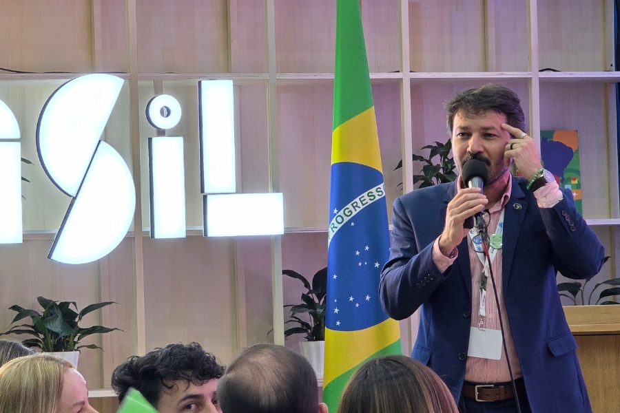 Homem de terno azul fala ao microfone enquanto aponta a mão para a cabeça. Ao fundo, a bandeira do Brasil e a estrutura com o letreiro do país.