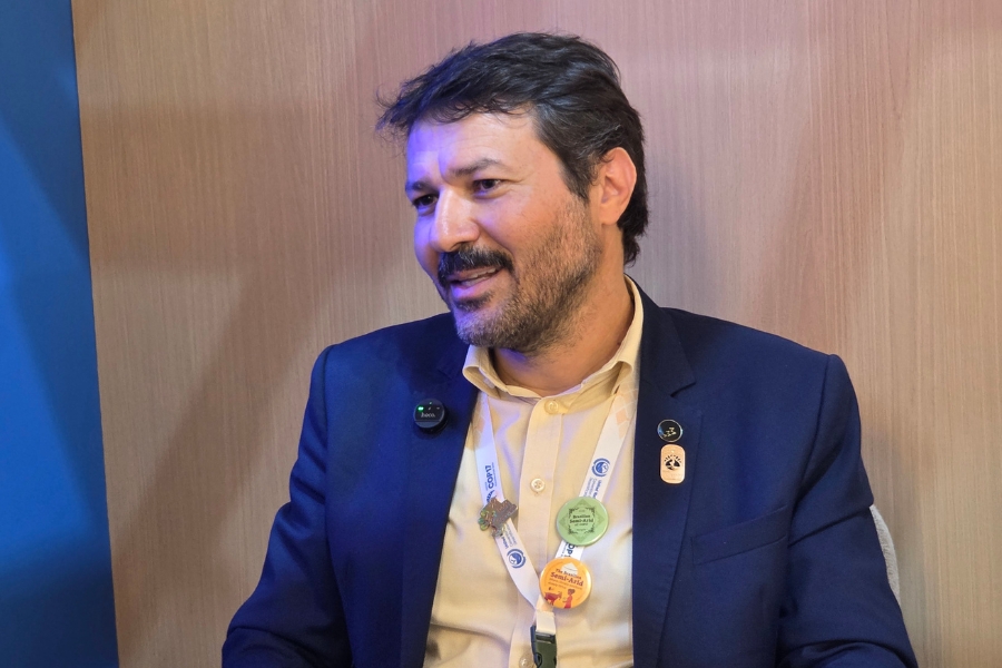 Foto de plano médio de um homem de meia-idade olhando ligeiramente para a esquerda e com a boca levemente aberta, indicando que está falando. Ele tem cabelo curto e bagunçado, de cor castanha escura e preta, uma barba cheia e ligeiramente grisalha, além de bigode, e pele em tons médios, apresentando algumas linhas de expressão sutis ao redor dos olhos e na testa. O homem veste um terno azul-marinho sobre uma camisa social bege clara, adornada com vários pins. Na lapela direita do terno, há um pin circular verde-limão e um pin dourado em forma de pino com o design de um rosto. Ele usa uma credencial branca pendurada no pescoço por um cordão branco com o texto "COP17" visível em preto. A credencial está posicionada acima de um pin dourado circular com um design preto e um pin circular laranja maior que apresenta as palavras "THE BRAZILIAN SEMI-ARID" em letras pretas e o design de um touro. Na altura do peito, do lado esquerdo, há também um microfone de lapela preto preso ao terno. O fundo é composto por uma parede de madeira marrom claro, com uma seção azul visível no canto superior esquerdo