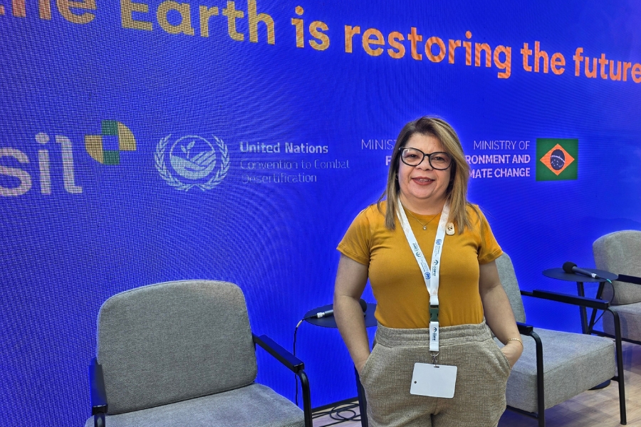 Fotografia em plano médio de uma mulher sorridente em um evento governamental e ambiental. Ela possui pele clara, cabelos loiros liso-ondulados na altura dos ombros, usa óculos de armação escura e arredondada, camiseta mostarda de mangas curtas e calça de tecido cinza-mescla com cós trabalhado, mantendo as mãos nos bolsos. No pescoço, carrega uma fita de credencial branca com um crachá de identificação em destaque na cintura. Ela está em pé entre poltronas cinza, posicionada à frente de uma grande tela azul onde se lê parcialmente o slogan em letras alaranjadas "is restoring the future" no topo, além dos logotipos do Brasil, da Convenção das Nações Unidas para o Combate à Desertificação (UNCCD) e da bandeira brasileira