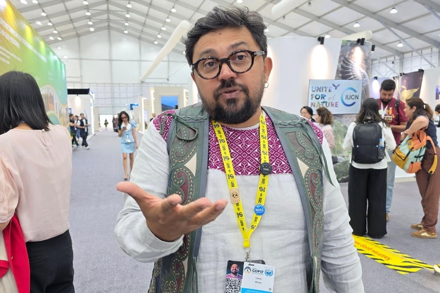 Homem de óculos, barba e veste bordada gesticula com a mão aberta em um grande galpão de eventos, com credencial do evento pendurada no pescoço.