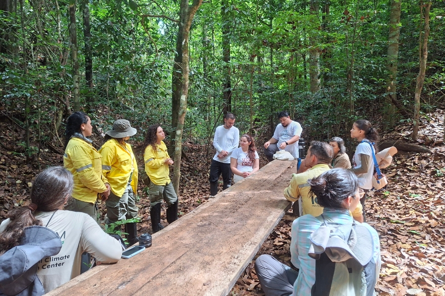 Um grupo de cerca de dez pessoas está reunido ao redor de uma mesa longa e rústica de madeira no meio de uma floresta densa. À esquerda, três integrantes usam uniformes amarelos de brigadistas ou agentes ambientais e calças escuras, enquanto permanecem de pé em conversa com os demais. Outras pessoas estão sentadas no chão ou em pequenos bancos em volta da grande bancada de madeira, vestindo roupas casuais de campo em tons claros e neutros. Ao fundo e nas laterais, a vegetação é composta por árvores altas e folhagens verdes vibrantes, com o chão totalmente coberto por folhas secas marrons sob a iluminação natural da mata