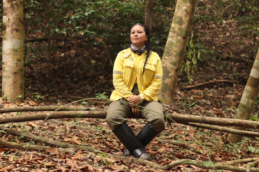 Uma mulher parda com cabelo escuro e algumas mechas cinzentas, vestindo uma camisa de uniforme amarela vibrante com tiras refletivas e o logotipo do ICMBio em verde, uma calça de carga verde-oliva e botas de borracha pretas altas, senta-se de pernas cruzadas em um tronco horizontal, olhando pensativamente para o lado em uma floresta densa com solo coberto de folhas secas e troncos de árvores. Ela tem uma bandana cinza no pescoço e uma trança no cabelo, com as mãos cruzadas no colo. Árvores altas de casca grossa emolduram a cena, com vegetação de sub-bosque verde escuro ao fundo. A iluminação é difusa