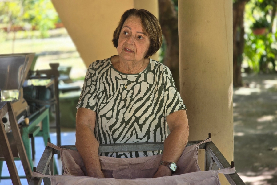Mulher idosa com blusa estampada de padrão abstrato olhada para o lado em ambiente coberto com pilares.