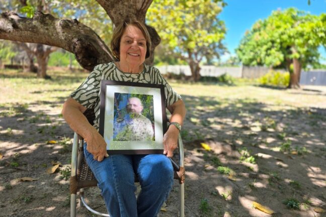 Mulher idosa sentada ao ar livre segurando um quadro com o retrato de outra mulher, sob a sombra de uma árvore.