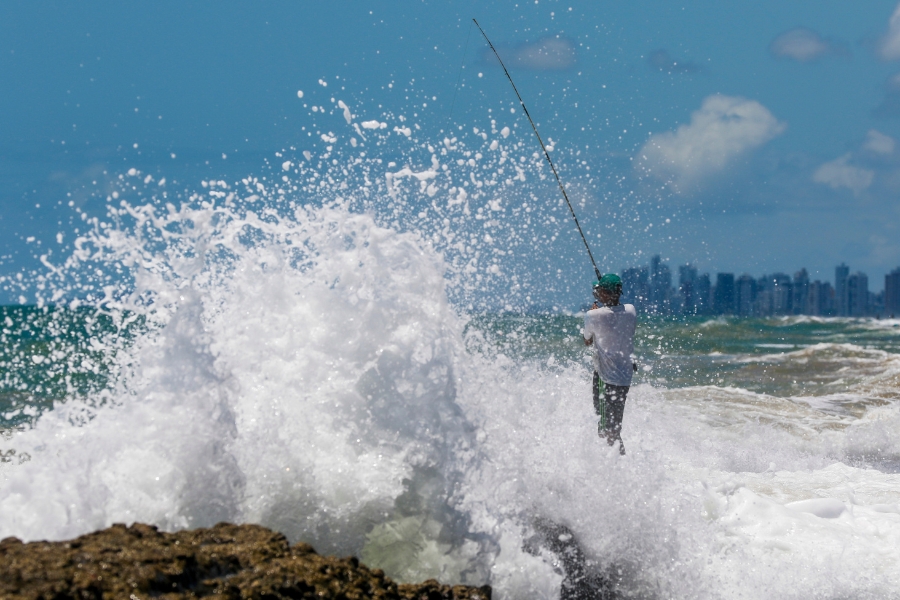 Fotografia de nível médio e médio-ângulo em cores mostra um pescador em pé no oceano, de costas para a câmera, enfrentando o spray do mar, com a orla da cidade à distância. O pescador está à direita do centro e está voltado para longe do ponto de vista. Ele tem pele de tom de azeitona, veste uma camiseta branca e shorts pretos. Ele está segurando uma vara de pesca longa com ambas as mãos, a vara diagonalmente para cima e para a esquerda. Ele tem o cabelo curto e preto e um chapéu de beisebol verde virado para trás. O pescador está imerso em água branca espumante e borrifada até a cintura. Uma onda enorme e turva com uma crista esbranquiçada sobe e cai no centro-esquerda. O spray da onda sobe alto no ar e à direita. O oceano é turvo e marrom no primeiro plano, verde-turquesa no meio e azul-esverdeado à distância. O horizonte é claro e nítido. A orla da cidade é uma linha de prédios altos e estreitos na distância no canto superior direito. O céu é azul-claro com nuvens brancas e difusas. O primeiro plano é composto por pedras marrom-claras no canto inferior esquerdo. A luz do sol vem da direita, lançando sombras suaves na cena