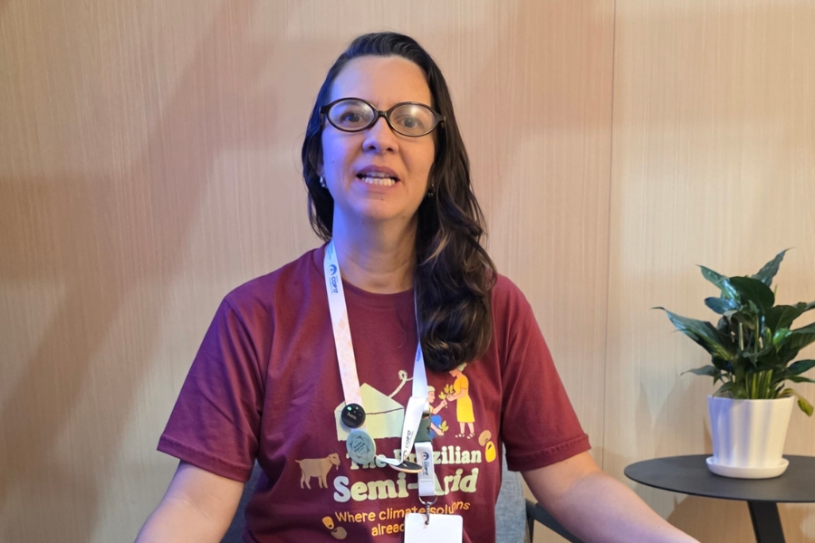 Uma mulher sorridente com cabelos escuros e ondulados está sentada e olha diretamente para a câmera, com os lábios levemente afastados como se estivesse no meio de uma frase. Ela usa óculos redondos de acetato castanho e uma camiseta marrom-avermelhada com a estampa de um bode, uma pessoa e um texto em inglês e branco que diz "The Brazilian Semi-Arid Where climate solutions already". Um cordão de crachá de identificação branco com letras "COP17" repetidas, está pendurado em seu pescoço com alguns botons nele. O crachá esta virado. No canto direito inferior, uma planta verde com folhas largas está plantada em um vaso de cerâmica branca, e tudo está posicionado em uma superfície de madeira marrom claro, com uma seção azul visível no canto superior esquerdo