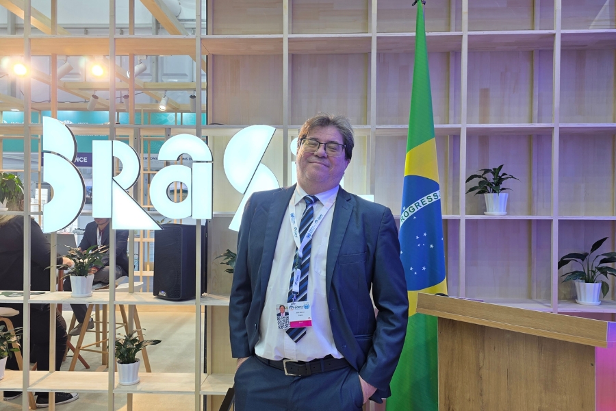 Homem de terno, gravata e óculos posa sorrindo em pé, com as mãos nos bolsos, em frente a uma estante de madeira, letreiro "Brasil" e a bandeira do país.