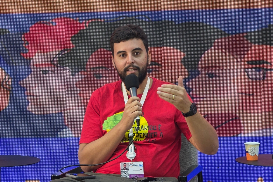 Homem de camiseta vermelha discursa com microfone em mãos. Ao fundo, painel digital com ilustração de rostos em perfil.