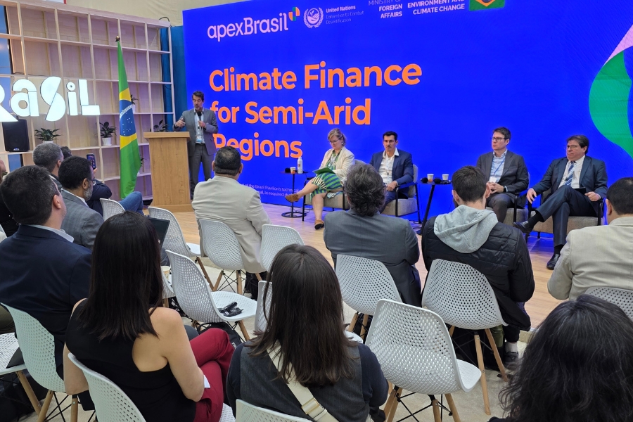 Painel com palestrantes sentados no palco. Homem ao púlpito fala diante do telão azul com a frase "Climate Finance for Semi-Arid Regions". Plateia assiste em primeiro plano.
