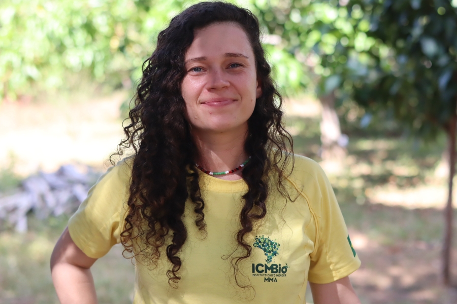 Um retrato em plano médio de uma mulher branca com cabelos longos e cacheados, de cor castanho-escura, que caem sobre seus ombros. Ela está de frente para a câmera, sorrindo e olhando diretamente para ela, em um ambiente externo com vegetação verde desfocada ao fundo. Ela veste uma camiseta amarela com gola redonda e mangas raglã que cobrem seus ombros, onde há uma pequena bandeira do Brasil na manga esquerda. No peito, está impresso o logotipo do ICMBio em verde: uma forma de folha ou árvore estilizada com a sigla ICMBio em letras maiúsculas e, abaixo, o texto INSTITUTO CHICO MENDES em letras maiúsculas menores e, mais abaixo, MMA em letras maiúsculas ainda menores. Ela usa um colar de contas coloridas que envolve o pescoço. O fundo desfocado mostra arbustos e árvores em tons de verde. A iluminação é natural e brilhante
