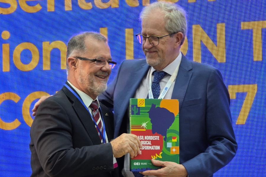 Dois homens de terno e óculos sorriem e se abraçam de lado enquanto seguram juntos uma publicação colorida. Ao fundo, vê-se parte de um telão azul com letras amarelas.