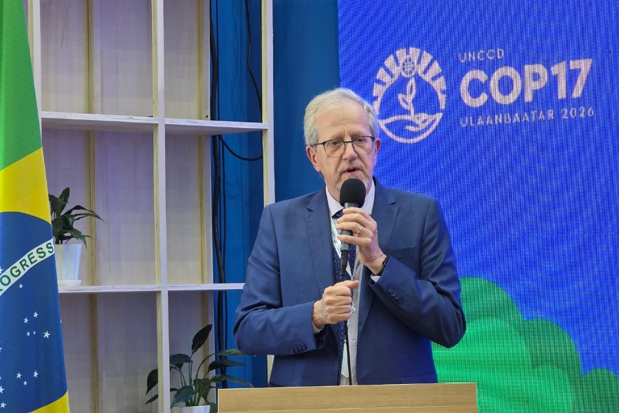 Homem de terno azul e óculos fala ao microfone em um púlpito de madeira. À esquerda está a bandeira do Brasil e, à direita, um telão azul com o logotipo da UNCCD COP17.