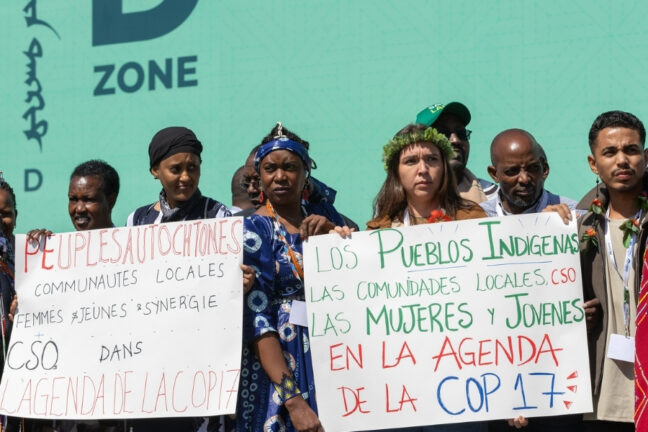 Grupo de ativistas segura dois cartazes reivindicando a inclusão de povos indígenas, mulheres e jovens na agenda da COP17, em francês e espanhol.