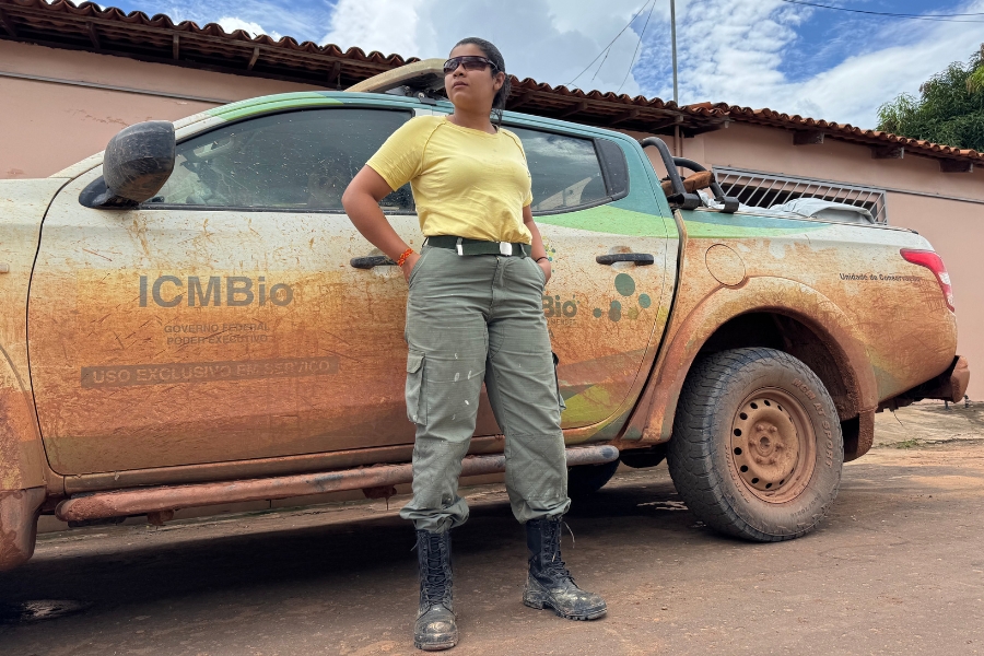 Uma foto de plano médio vertical mostra uma mulher em pé em frente a uma caminhonete de cabine dupla coberta de lama em uma estrada de terra sob um céu parcialmente nublado, vestindo uma camiseta amarela, calças cargo verde-oliva com manchas de lama branca e botas de combate pretas. A mulher, que está de frente para a câmera e com a mão direita no quadril, tem cabelos escuros presos e usa óculos de sol, olhando para longe. A caminhonete branca e verde, cujas portas laterais, roda dianteira e para-choque traseiro estão cobertos de lama seca, tem as palavras "ICMBio" e "UNIDADE DE CONSERVAÇÃO" impressas em letras maiúsculas verdes na porta dianteira e lateral, respectivamente. Uma placa de metal verde também é visível no para-lama traseiro, que parece dizer "SISTEMA NACIONAL DE UNIDADES DE CONSERVAÇÃO DA NATUREZA" em letras verdes. A roda dianteira e o pneu estão cobertos de lama. Ao fundo, uma casa rosa com telhado de telha marrom é visível