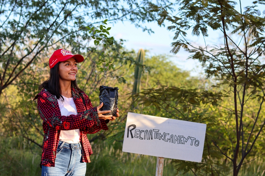Mulher sorridente com boné vermelho e camisa xadrez segura uma muda vegetal ao lado de uma placa com a palavra "RECAATINGAMENTO".