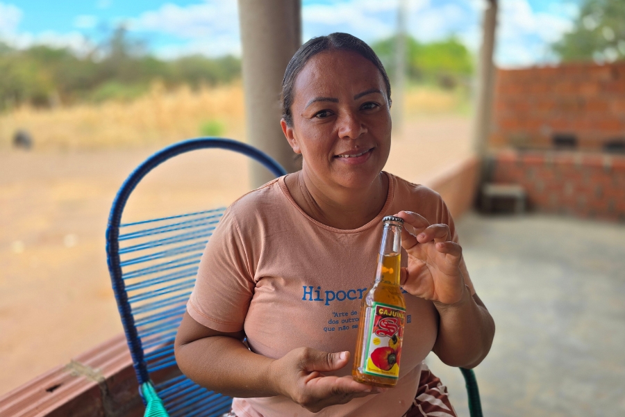 Mulher sorrindo segurando uma garrafa de cajuína em área externa. Veste camiseta rosa e está sentada em uma cadeira azul.