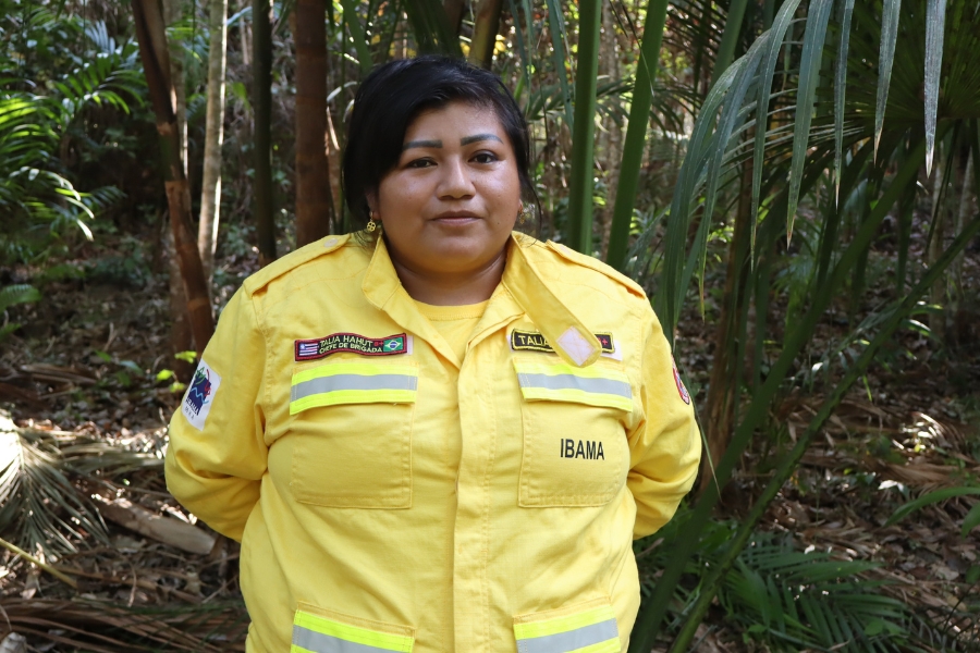 Fotografia em plano médio de uma mulher indígena no centro da imagem, posicionada de frente com os braços para trás e olhando serenamente para a câmera. Ela tem cabelos pretos e curtos, pele morena e usa uma camisa amarela de mangas longas da brigada do IBAMA, com faixas refletivas amarelas e cinzas no peito e na cintura, bolsos frontais e identificação bordada com seu nome e o texto "Chefe de Brigada". O cenário ao fundo é de floresta tropical, iluminada por luz natural que atravessa a copa das árvores, destacando folhagens verdes intensas, palmeiras e o solo coberto por folhas secas
