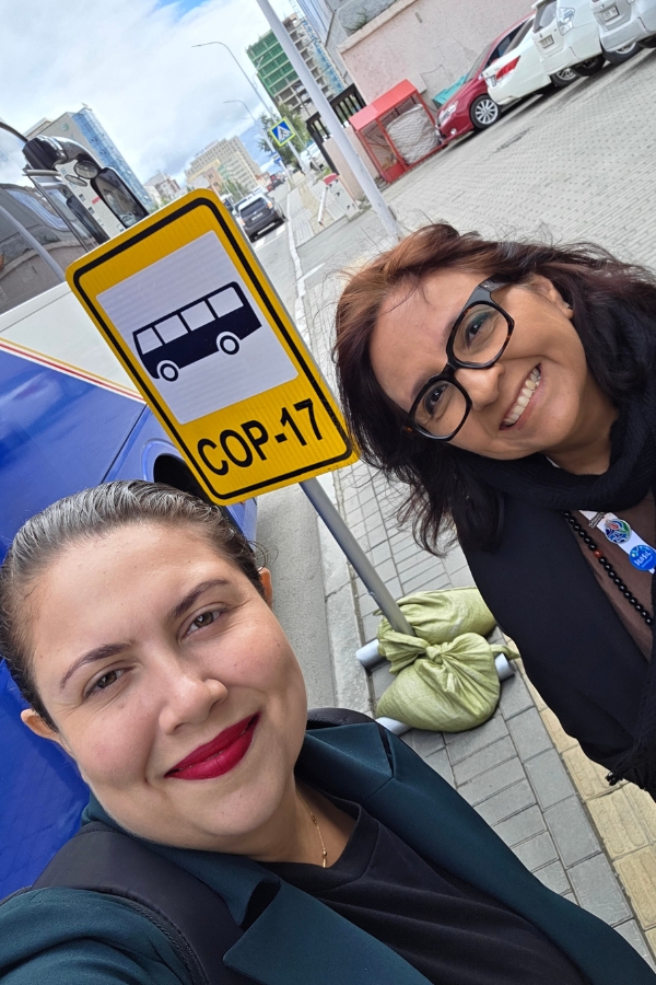 Duas mulheres tiram uma selfie sorrindo ao lado de uma placa amarela de sinalização do ônibus da COP-17 em uma rua da cidade.