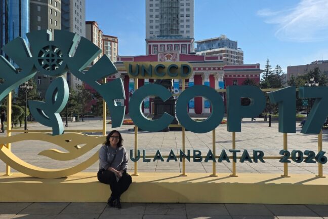 Uma mulher está sentada na estrutura do letreiro da "UNCCD COP17 Ulaanbaatar 2026" em uma praça.