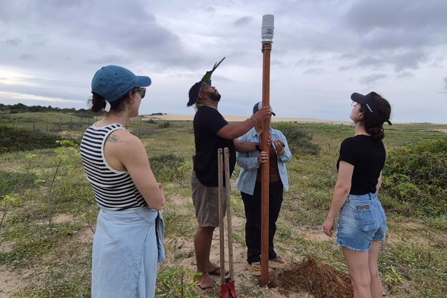 O grupo reunido ao redor do mesmo poste da estação meteorológica em campo aberto, observando o fixamento e o ajuste do equipamento no topo.