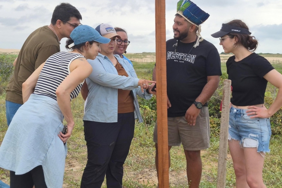 Registro em plano mais próximo do grupo reunido ao redor da haste de madeira. Uma pesquisadora aponta para a estrutura enquanto explica o funcionamento ao indígena Tremembé e demais presentes.