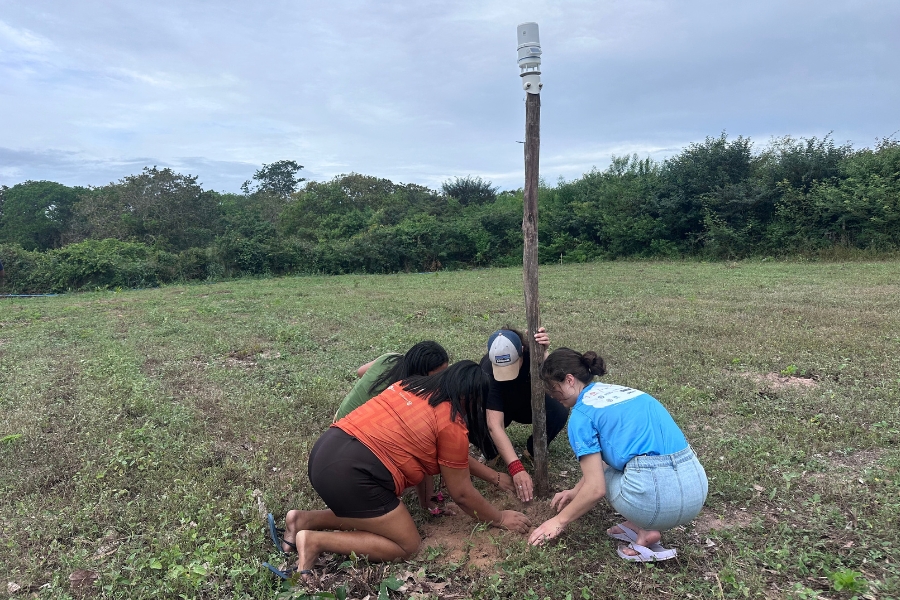 Grupo de quatro pessoas ajoelhadas na terra ao redor da base do poste da estação meteorológica para fixá-lo firmemente ao solo em campo aberto.