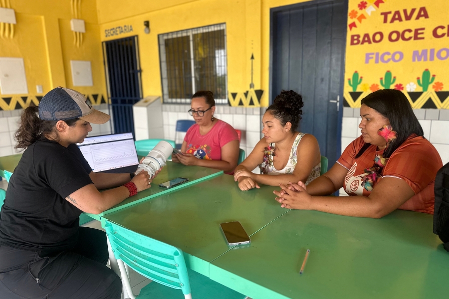 Reunião em ambiente escolar/comunitário ao redor de uma mesa verde. Uma pesquisadora com boné demonstra um sensor portátil e exibe dados na tela do notebook para três mulheres (uma delas vestindo colar/adorno tradicional).