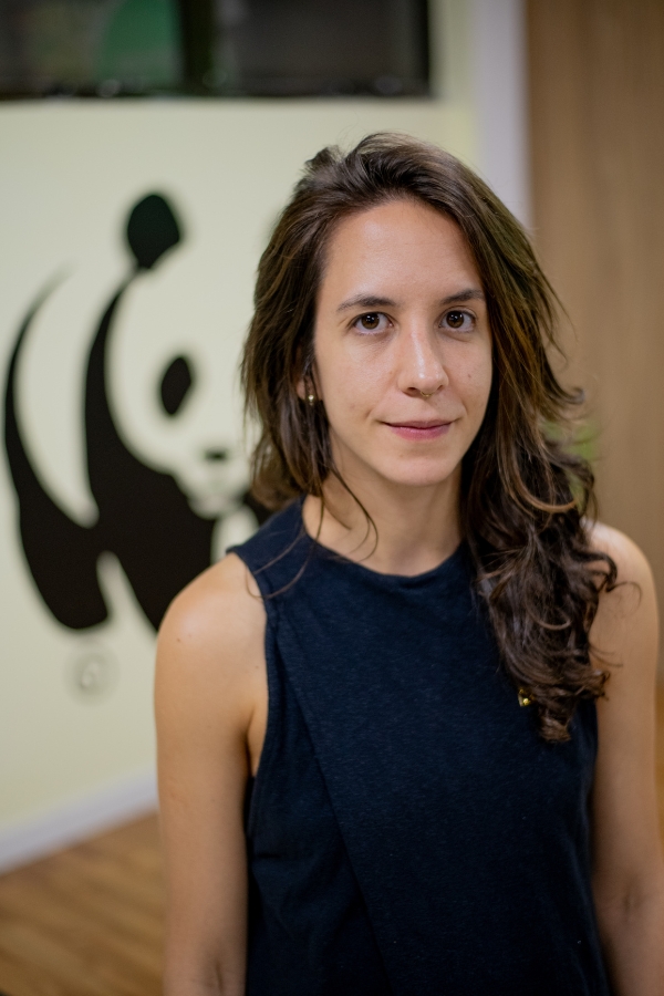 Mulher branca de cabelos castanhos e blusa preta sorrindo em um ambiente interno com o logotipo da WWF ao fundo.