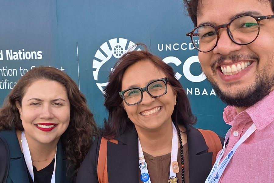 Três jornalistas, duas mulheres brancas e um homem branco, sorriem em uma selfie em frente ao painel do evento da UNCCD.