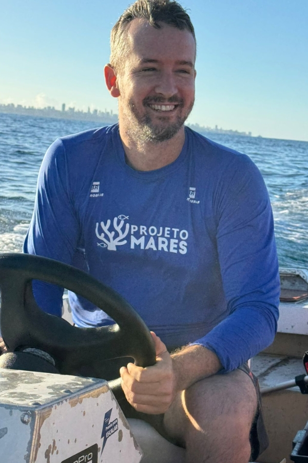 Homem sorridente vestindo camiseta do Projeto Mares conduz uma embarcação no mar.