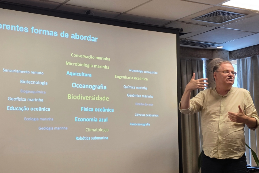 O pesquisador Roberto Christofoletti ministrando uma palestra ao lado de uma tela de projeção que lista diversas áreas de estudo sobre a conservação das ciências do mar (como Oceanografia, Biodiversidade, Conservação marinha, Economia azul, entre outras).