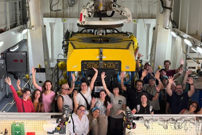 Grupo de pesquisadores e membros da equipe sorrindo e acenando em frente ao robô submarino (ROV - Remotely Operated Vehicle) SuBastian, de cor amarela, a bordo da embarcação.