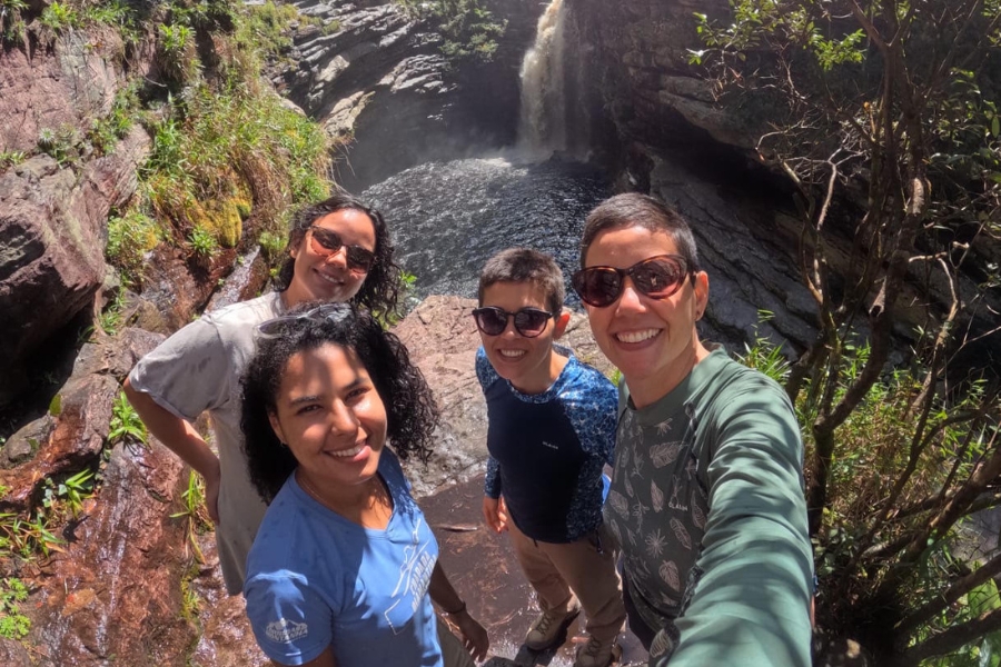 Quatro pessoas sorriem para uma selfie em um mirante rochoso com uma cachoeira e poção ao fundo.