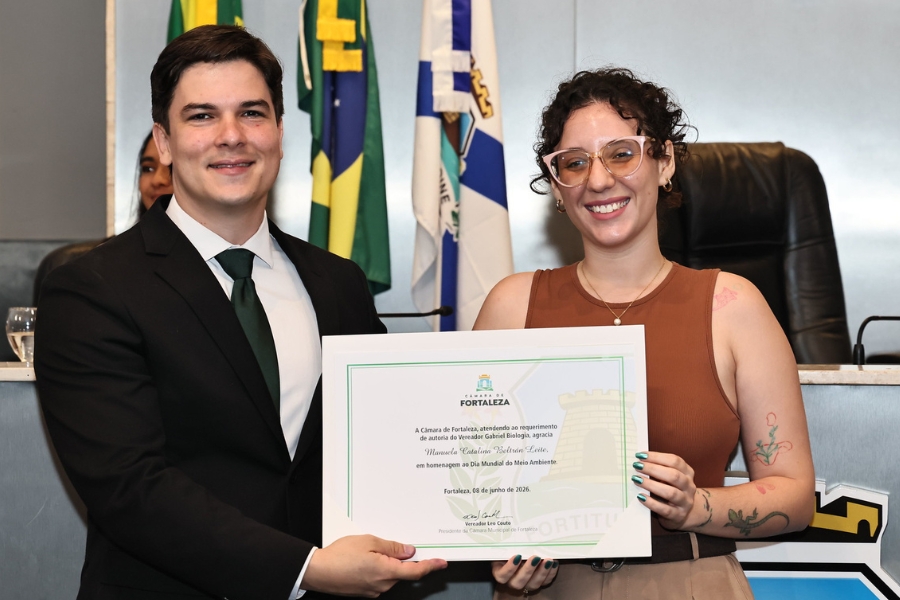 Homem de terno e gravata entrega um certificado de homenagem em sessão solene da Câmara Municipal de Fortaleza a uma mulher sorridente.