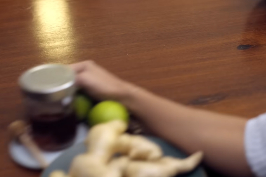 Imagem em desfoque mostrando uma mão alcançando ingredientes para remédio caseiro (pote com mel, limões e gengibre) dispostos sobre uma mesa de madeira.