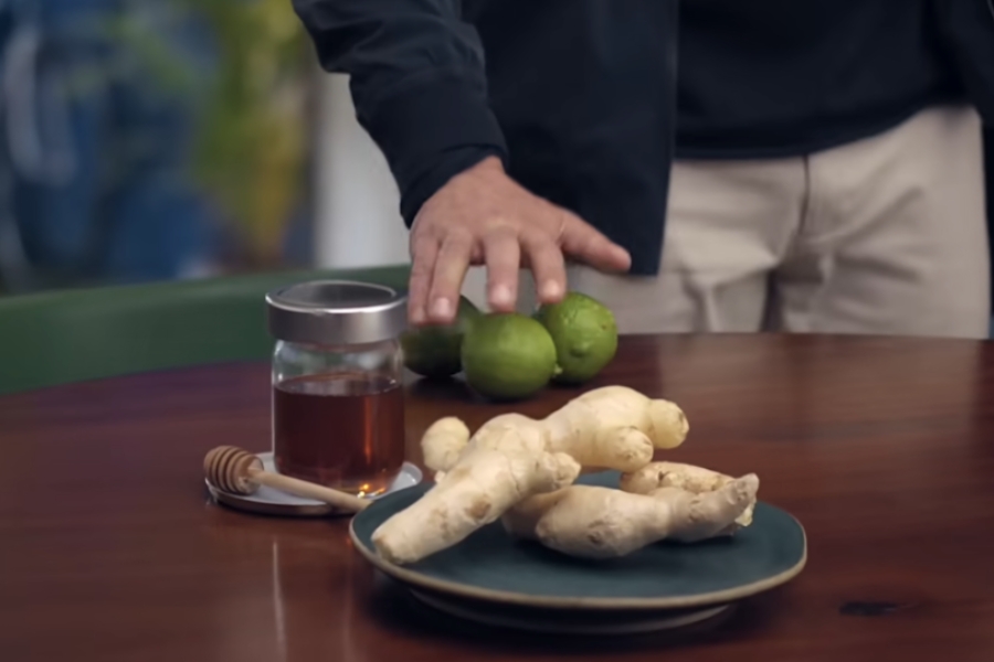 Uma pessoa aproximando a mão de ingredientes dispostos sobre a mesa, incluindo raízes de gengibre em um prato, limões e um frasco com mel.