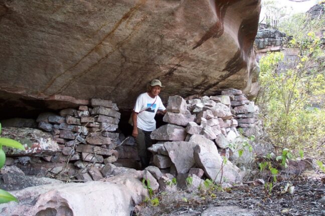 Homem de chapéu dentro de uma construção histórica de pedras sob uma enorme rocha saliente