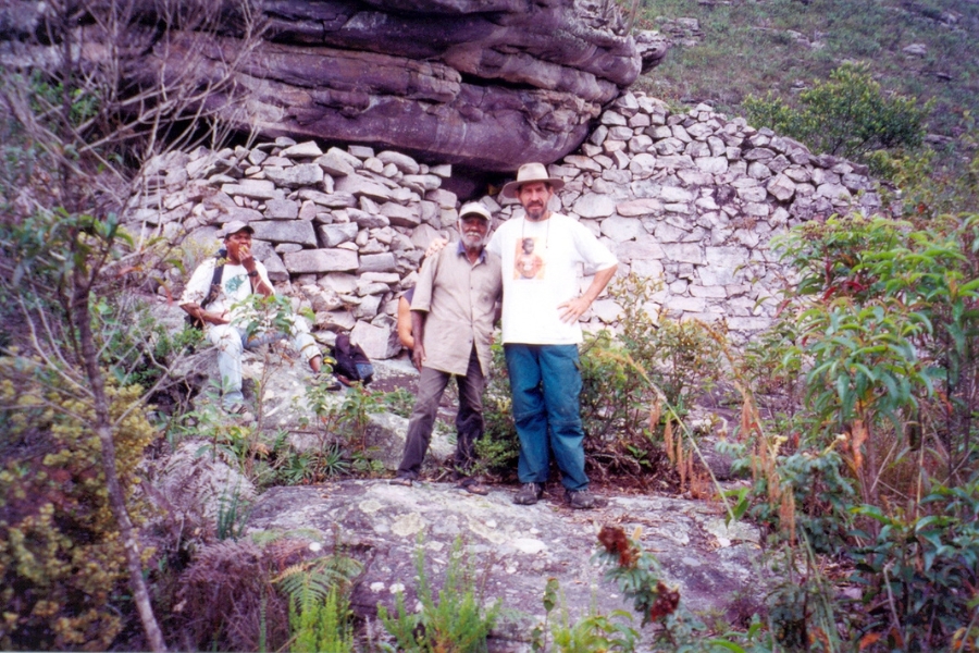 Dois homens em pé e um sentado junto a uma antiga parede de pedras empilhadas na base de um paredão rochoso.